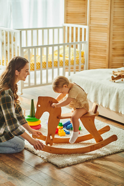 Een jonge moeder speelt vrolijk met haar peuterdochter op een traditioneel houten hobbelpaard, verbindt en creëert dierbare herinneringen. - Foto, afbeelding