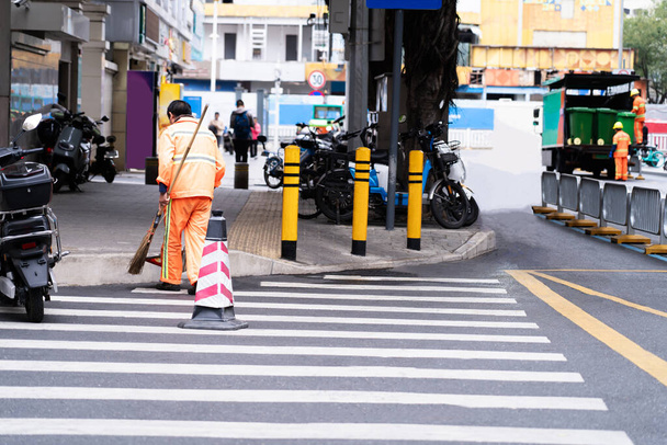 Ένας άνθρωπος σαρώνει τους δρόμους με μια σκούπα, καθαρούς δρόμους της πόλης Καθαρίστε την πόλη από τα σκουπίδια. Δημοτικοί υπάλληλοι με πορτοκαλί στολές σαρώνουν τους δρόμους. - Φωτογραφία, εικόνα