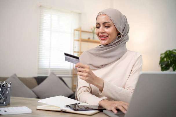 Jonge gelukkige moslim vrouw in hijab thuis met behulp van laptop winkelen online met creditcard terwijl zitten op het bureau. - Foto, afbeelding