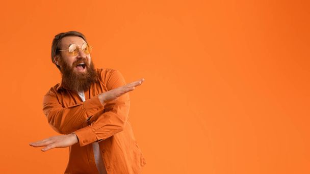 Red-haired man with beard dressed in bright shirt and sunglasses dances joyfully, crossing hands with excitement on orange studio background. Concept of positivity and cool party vibes. Panorama - 写真・画像