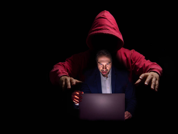 Una figura encapuchada manipula a un hombre de negocios que trabaja en su computadora portátil, simbolizando estafas en línea. - Foto, Imagen