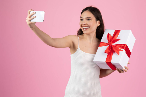 Beyaz elbiseli neşeli bir kadın pembe arka planda kırmızı kurdeleli büyük beyaz bir hediye kutusuyla selfie çeker. - Fotoğraf, Görsel