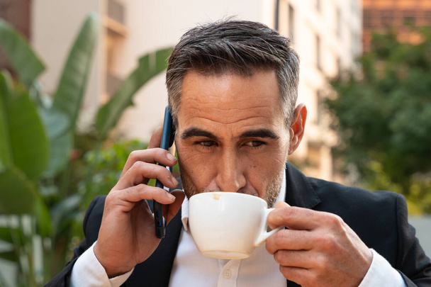  Mężczyzna w średnim wieku pijący kawę szczęśliwie rozmawiający z telefonem komórkowym w pracy. - Zdjęcie, obraz