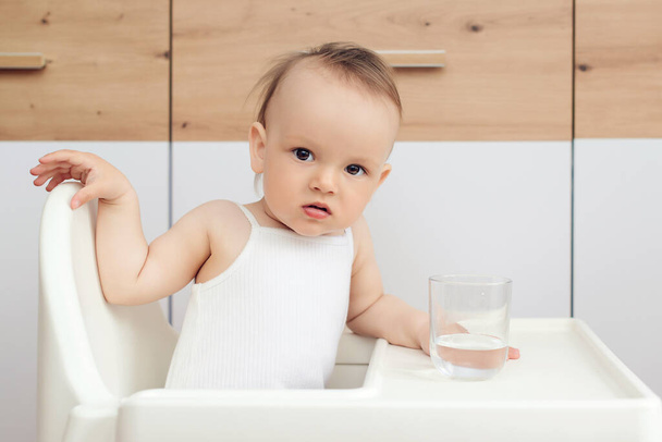 Doce menina segurando vidro e água potável. Feliz bebê sentado em uma cadeira de bebê na cozinha. Criança bebe água. - Foto, Imagem
