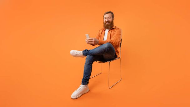 Hombre con el pelo rojo y la barba utiliza el teléfono móvil sonriendo con satisfacción mientras está sentado en la silla, navegar por Internet y mensajes de texto a través del dispositivo, estudio de tiro sobre fondo naranja. Panorama, espacio vacío - Foto, imagen