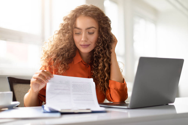 Een jonge vrouw met krullend haar is gericht als ze beoordeelt documenten aan haar bureau met een laptop in de buurt op kantoor - Foto, afbeelding