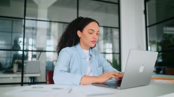 Motivovaný soustředěný nádherný brazilský nebo hispánský kudrnatý žena v pastelově modré košili, zaměstnanec společnosti, sedí v moderní kanceláři u pracovního stolu, pracuje na notebooku, soustředěný dělá poznámky do notebooku - Záběry, video