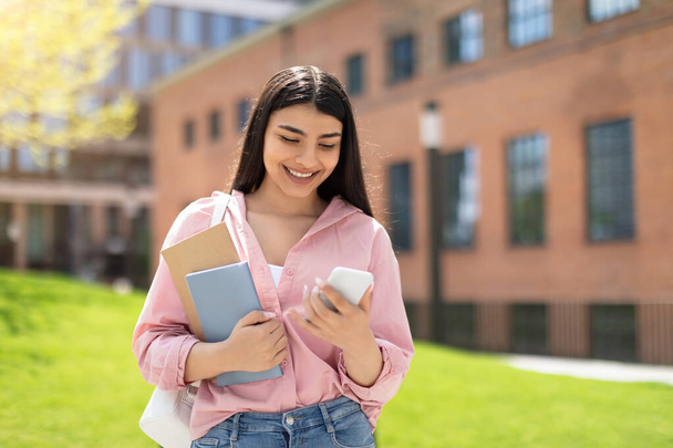 Młoda dziewczyna z notatnikami uśmiecha się, gdy patrzy na smartfona z budynkiem kampusu w tle - Zdjęcie, obraz