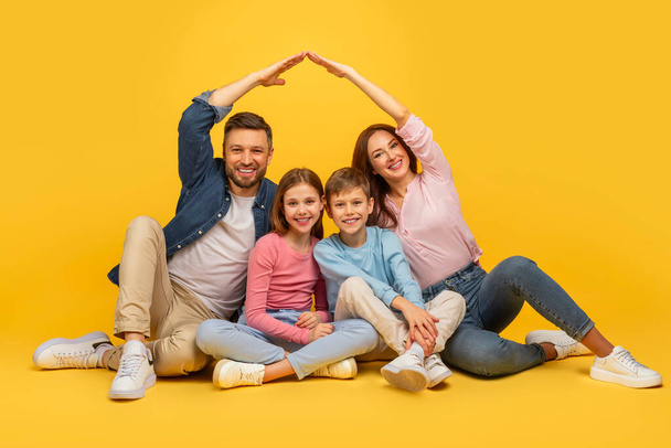 床に座っている家族,両親の間の子供,明るい黄色の背景に一緒に手で家の屋根の形を作る - 写真・画像