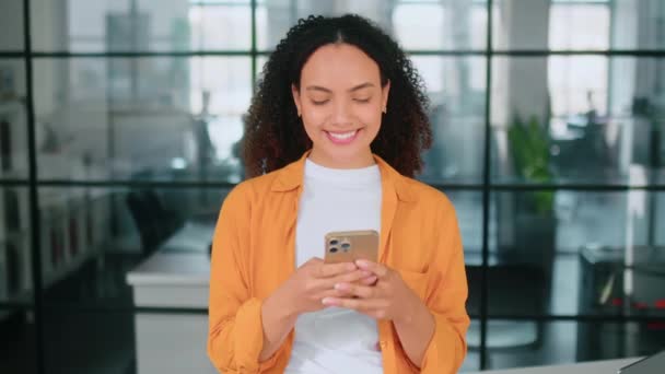 Een mooie positieve Braziliaanse of Spaanse jonge vrouw met krullend haar in een elegante jas, staat in een moderne kantoorruimte, gebruikt haar smartphone, berichten met vrienden op sociale netwerken, glimlacht - Video