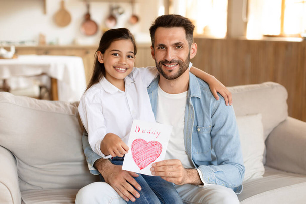 Usmívající se otec a jeho mladá dcera sedí na gauči s ručně dělanou tatínkovou kartou, vyjadřující lásku a radost - Fotografie, Obrázek