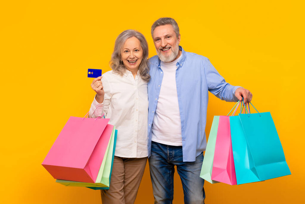 Happy senior couple showcases online shopping joy with credit card and colorful shopper bags, standing over yellow studio backdrop, smiling to camera. Concept of Black Friday sales - Photo, Image