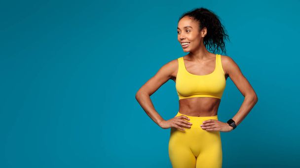 Mujer afroamericana atlética confiada posa en ropa deportiva amarilla sobre un fondo azul vibrante, mostrando fitness y salud - Foto, imagen