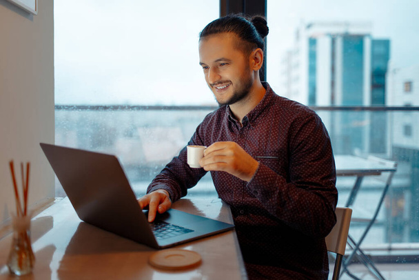 Portret van een knappe man die werkt op een laptop, typt op een toetsenbord terwijl hij een klein kopje espresso koffie vasthoudt op de achtergrond van panoramische ramen. - Foto, afbeelding