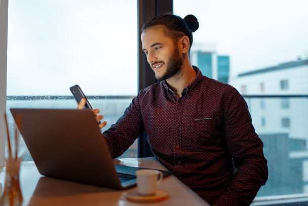 Portrait of handsome smiling man looking in smartphone while working at laptop, background of panoramic windows. - Photo, Image
