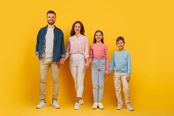 Affectionate family standing and smiling together on a yellow backdrop, father mother and kids holding hands - Photo, Image