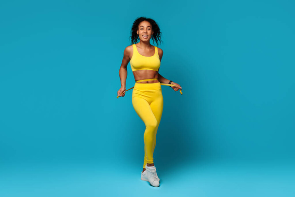 A happy, energetic African American woman in a yellow sports bra and leggings measuring waist against a vibrant blue background, representing health and vitality - Photo, Image