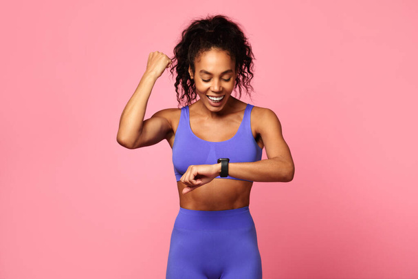Una alegre mujer afroamericana de fitness en un sujetador deportivo azul y polainas revisando su reloj inteligente sobre un fondo rosa - Foto, Imagen