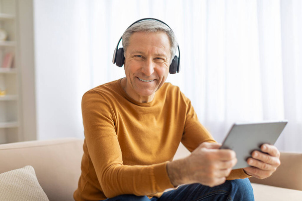 Senior man genieten van een digitale tablet en luisteren naar audio via een koptelefoon met een glimlach, interieur - Foto, afbeelding