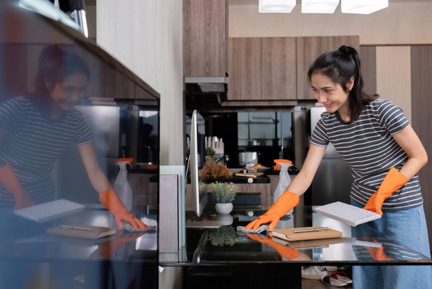 Krásná mladá hospodyně nosí oranžové rukavice a používá hadřík k čištění stolu a obývacího pokoje.. - Fotografie, Obrázek