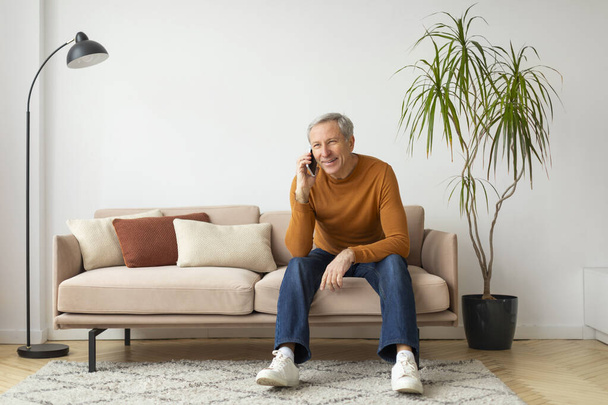 Cheerful mature man is seated on a beige couch in a well-lit room talking on a smartphone, copy space - Photo, Image