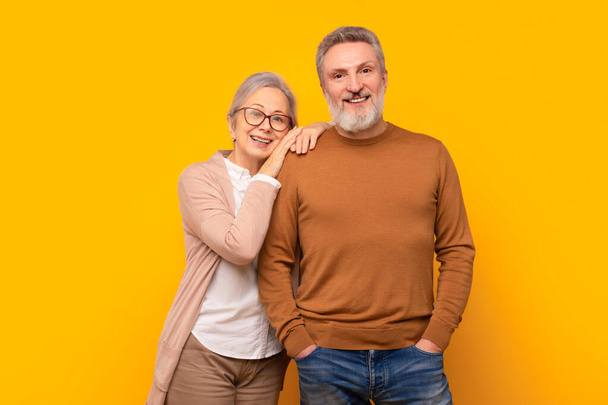 Ευτυχισμένο ζευγάρι Καυκάσιων ηλικιωμένων αγκαλιάζεται σε κίτρινο φόντο στούντιο, ακτινοβολώντας αγάπη και ζεστασιά του μακροχρόνιου γάμου. Έννοια της ώριμης στοργής και της διαρκούς συντροφικότητας - Φωτογραφία, εικόνα