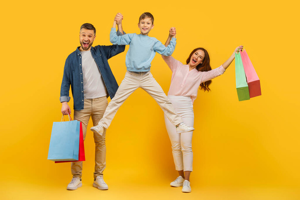 Eine energiegeladene und fröhliche Familie mit Einkaufstüten, die auf einem leuchtend gelben Hintergrund springen und feiern - Foto, Bild