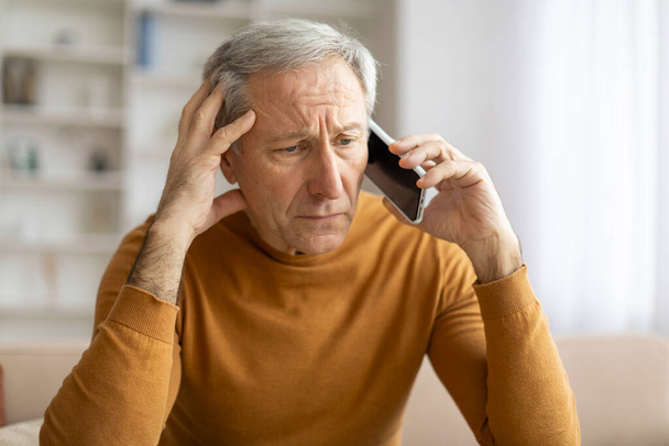 Homme mûr préoccupé tient un téléphone à son oreille, semble inquiet ou concentré alors que dans un cadre domestique - Photo, image