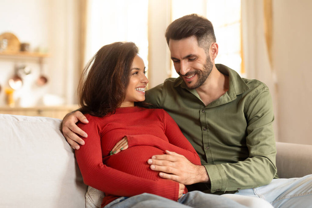Una pareja expectante comparte un momento tierno en el sofá, con el enfoque en el vientre de las mujeres embarazadas - Foto, Imagen