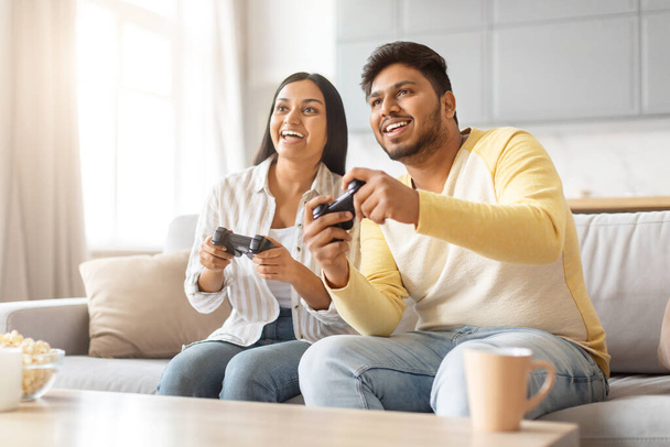 Indiano homem e mulher estão sentados em um sofá, imersos em jogar um jogo de vídeo juntos. - Foto, Imagem