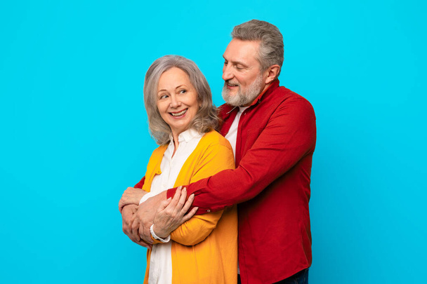 Glücklich liebendes Senioren-Paar, das sich vor blauem Hintergrund im Studio liebevoll umarmt und Liebe und Verbundenheit zwischen reifen Partnern zeigt. Grauhaariger Ehemann umarmt seine Frau zärtlich von hinten - Foto, Bild