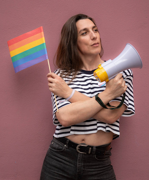 Транссексуальная женщина смотрит в сторону и держит в руках радужный флаг и мегафон в поддержку ЛГБТК-сообщества как активиста. - Фото, изображение