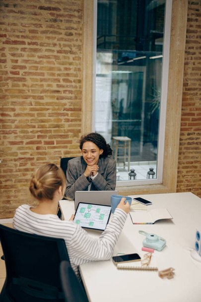 Two smiling female freelancers talking during work on laptop while sitting in cozy coworking - Foto, Imagem
