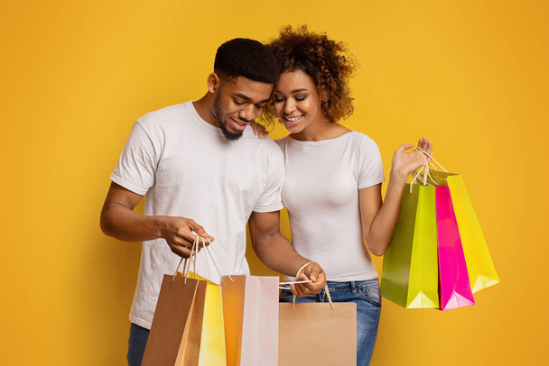 Sonriendo joven pareja afroamericana mirando en bolsas de compras con alegría contra un fondo amarillo vibrante, lo que sugiere una exitosa juerga de compras - Foto, imagen