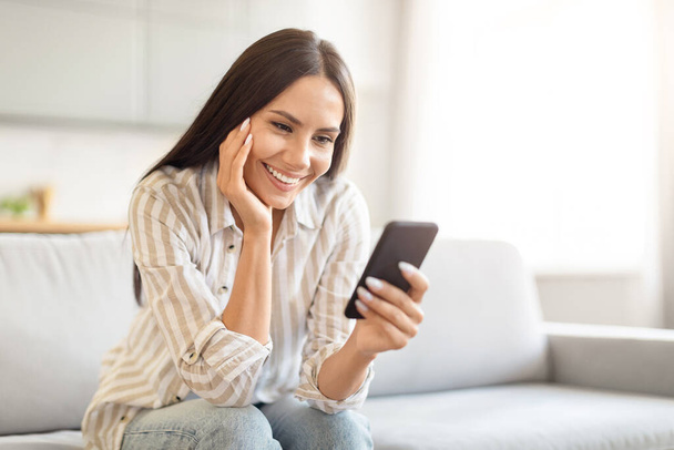 Mujer con una sonrisa contenta mirando el teléfono inteligente, sentado en un sofá de la sala de estar luminoso y aireado, espacio para copiar - Foto, Imagen