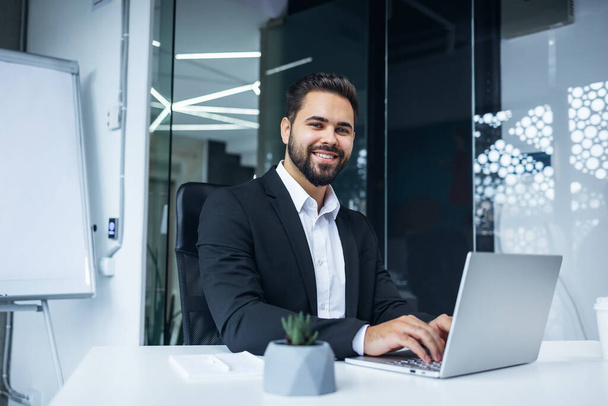 Modern Office Portrait of Stylish Hispanic Businessman Work on Laptop. makes Data Analysis, Looks at Camera and Smiles. Entrepreneur Works on e-Commerce Startup Project - Photo, Image