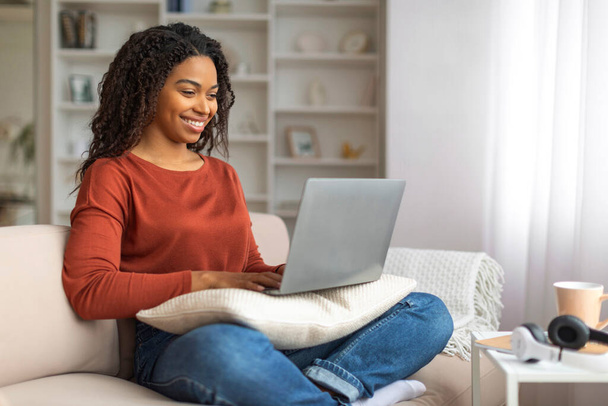Önünde dizüstü bilgisayarı açık bir şekilde kanepede oturan mutlu siyahi kadın, ekranda daktilo ediyor ve geziniyor, evden internet üzerinden çalışıyor, bedava boşluk. - Fotoğraf, Görsel