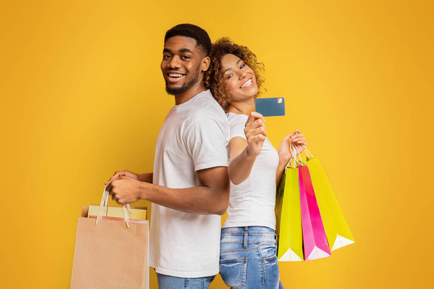 Nos encanta nuestro banco feliz pareja afroamericana milenaria con bolsas de compras y tarjeta de crédito, fondo naranja
 - Foto, imagen