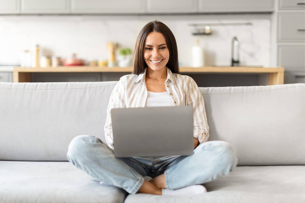 Με τα πόδια σταυρωμένα στον καναπέ, μια γυναίκα χαμογελά καθώς χρησιμοποιεί το laptop της σε ένα χαλαρό σπιτικό περιβάλλον - Φωτογραφία, εικόνα