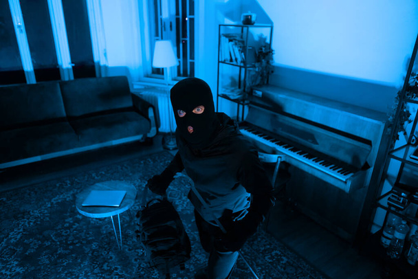 Ladrão mascarado com uma mochila inspeciona um ambiente de sala de estar, planejando seu próximo movimento com cuidado - Foto, Imagem