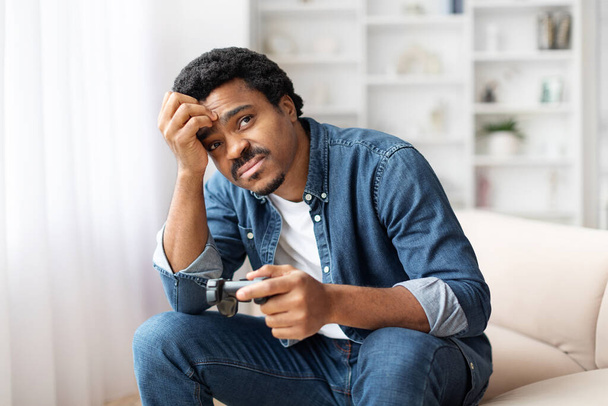 Genç siyah bir adam kanepede oturuyor, elinde joystick tutarken endişeli görünüyor, evinde video oyunları oynuyor ve kafaya dokunuyor. - Fotoğraf, Görsel