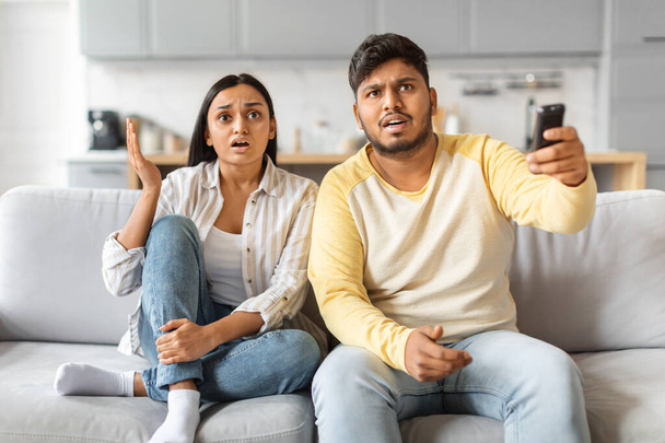 Para Indian siedząca na kanapie wykazuje szok i napięcie podczas oglądania razem telewizji, młoda para emocjonalnie reaguje na treść - Zdjęcie, obraz