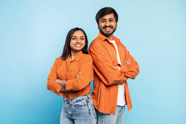 Dwóch młodych Indian mężczyzna i kobieta w pomarańczowych koszulach pozowanie z ramionami skrzyżowane, uśmiechając się na niebieskim tle - Zdjęcie, obraz