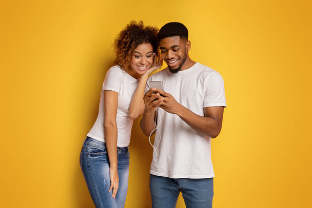 Young African American man and woman enjoy music together using one pair of earbuds against a yellow background - Fotó, kép