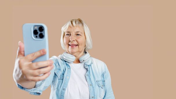 Positif femme âgée heureuse portant des vêtements décontractés pose pour selfie tenant smartphone dans une main. Vieillesse active concept - Photo, image