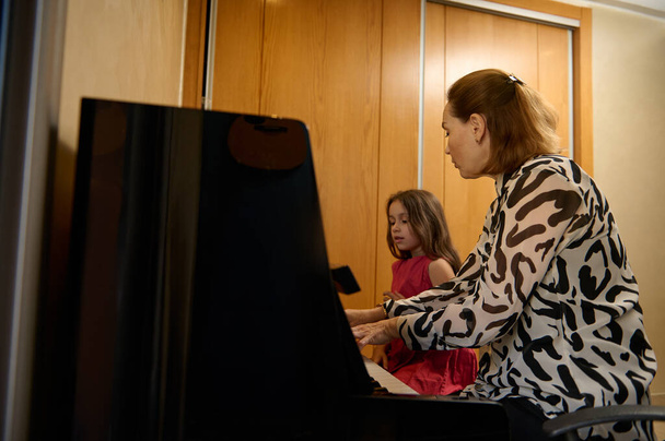 Автентичний бічний портрет кавказької молодої дорослої жінки, вчителя музики з учнем, що сидить на старовинному дерев'яному піаніно вдома, дає музичний урок. Мати і дочка грають на піаніно разом - Фото, зображення