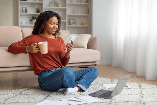 Usmívající se černoška sedící se zkříženýma nohama na podlaze s šálkem kávy a smartphonem, africká americká žena obklopená dokumenty a notebookem. - Fotografie, Obrázek