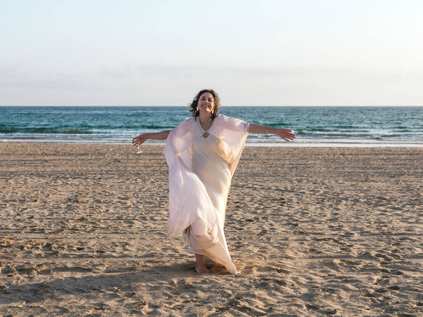 Une femme en blanc, les bras ouverts sur une plage sereine, face à l'océan. - Photo, image