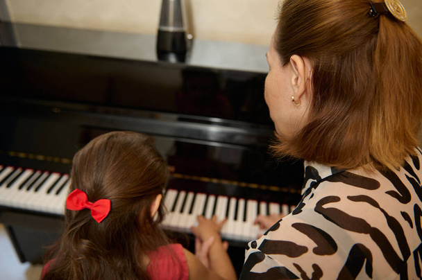 女性ミュージシャンピアニストのリアビューは,個々の音楽クラスで小さな女子生徒にピアノのレッスンを与えます. 教育,子供のエンターテイメント,芸術的な発展。 趣味とレジャー - 写真・画像