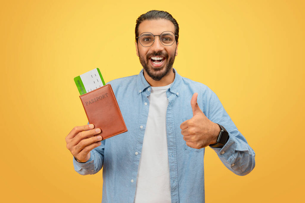 Joyful indian man shows approval with passport and boarding passes on yellow, signaling positive travel experience - Photo, Image